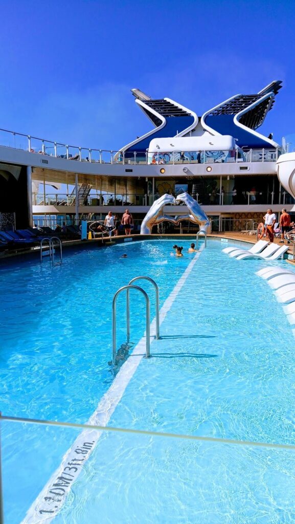 The main pool area on the Celebrity Apex cruise ship, featuring clear blue water and sun loungers partially submerged for relaxation. A modern sculpture of two hands forming a heart shape is visible in the background, along with the ship's distinctive logo and upper deck. Passengers are enjoying the sunny weather by the pool.