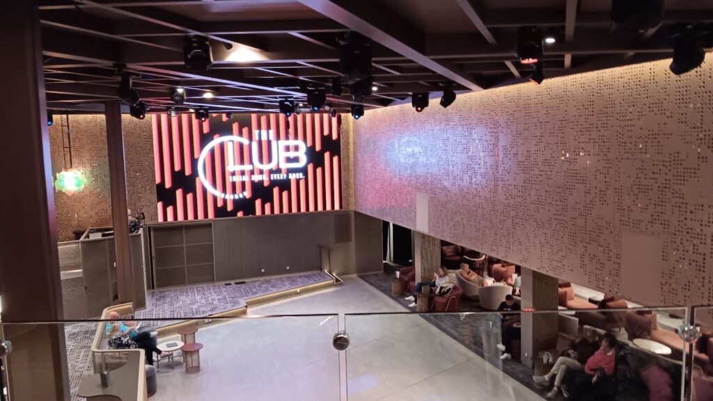 The Club on the Celebrity Apex cruise ship, featuring a modern, spacious interior with comfortable seating areas and a large digital screen displaying "The Club" logo. The venue offers a social space for passengers to relax and enjoy entertainment, with a stylish and contemporary design.
