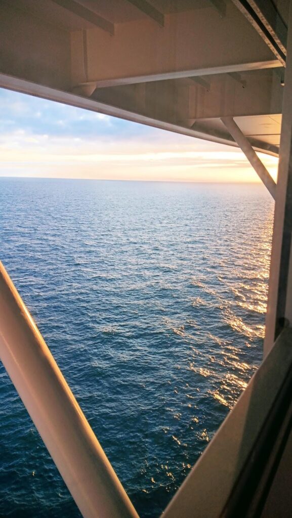 A view of the open ocean at sunset from a window on the Celebrity Apex cruise ship. The sky is painted with warm hues as the sun sets, reflecting off the calm sea, framed by the ship's structure.
