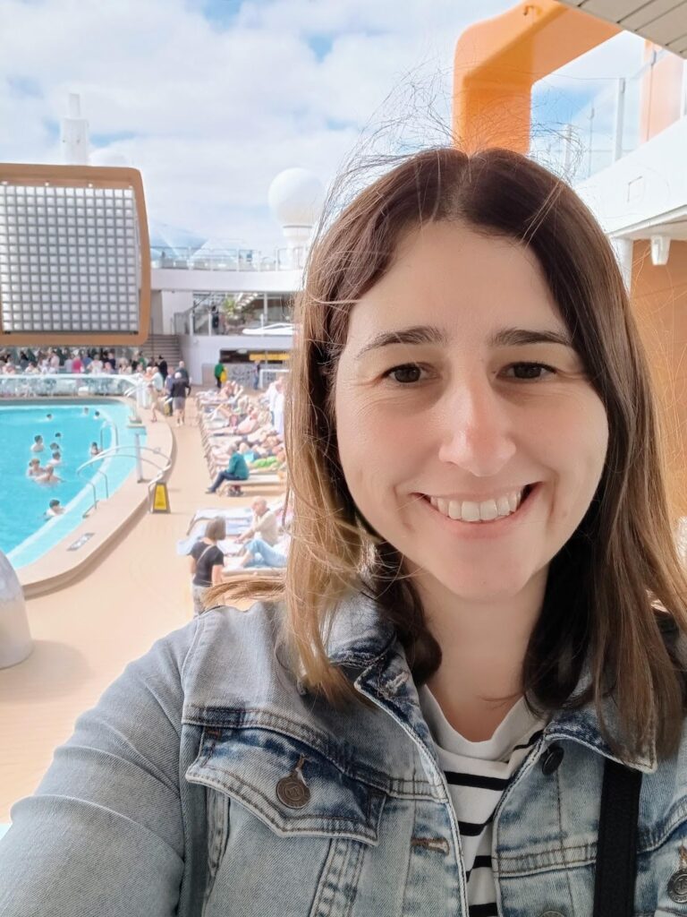 A woman smiling and taking a selfie on the deck of the Celebrity Apex cruise ship, with the pool area and numerous people lounging in the background. The weather appears to be pleasant, with a partly cloudy sky.