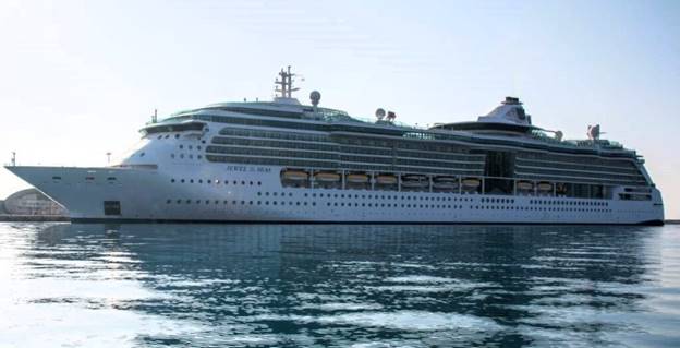 The serene waters mirror the Royal Caribbean's Jewel of the Seas, a sophisticated and sleek cruise ship docked in a peaceful harbor. The ship's design, with its rows of balconies and expansive glass windows, offers a glimpse into the elegant experiences awaiting guests aboard.