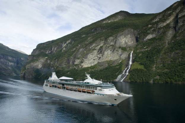 The 'Vision of the Seas' cruise ship glides through calm waters in a fjord, flanked by steep green cliffs and a cascading waterfall, under a partly cloudy sky, evoking a sense of adventure in the Norwegian landscape.