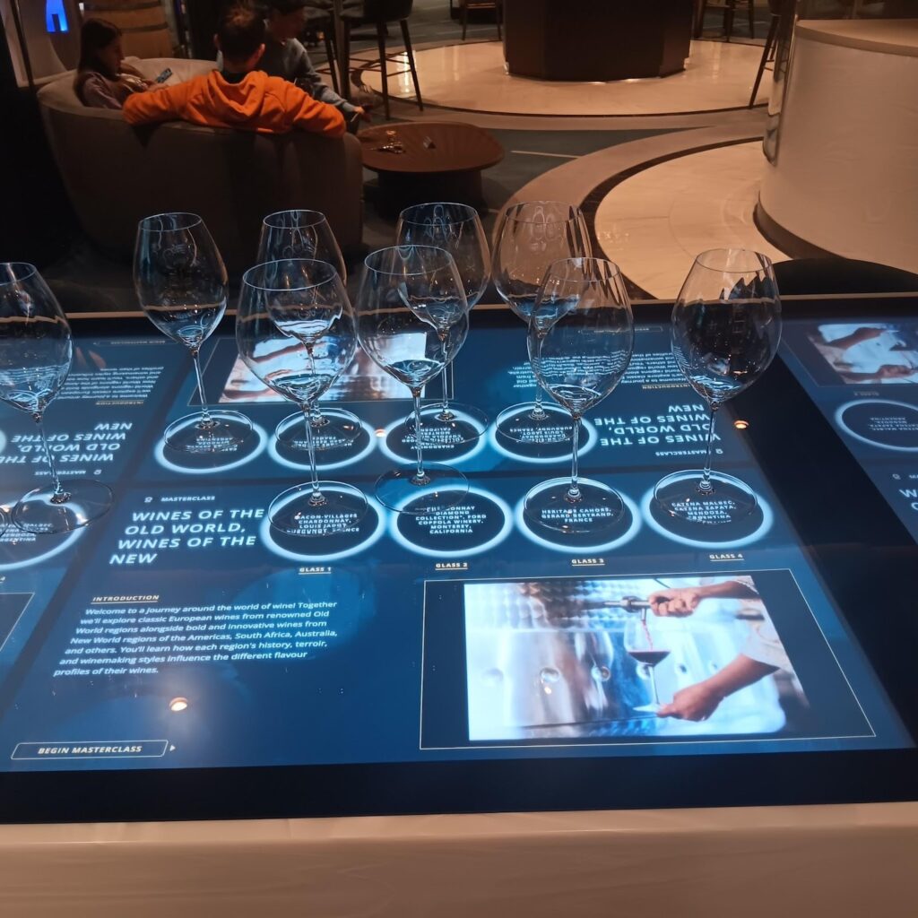  Interactive digital display for a wine masterclass, with empty wine glasses lined up above descriptions of Old World and New World wines. The display is in a social setting with individuals seated in the background.
