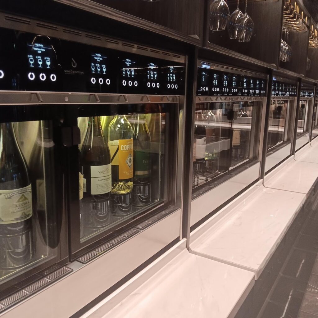 Interactive digital display for a wine masterclass, with empty wine glasses lined up above descriptions of Old World and New World wines. The display is in a social setting with individuals seated in the background.
