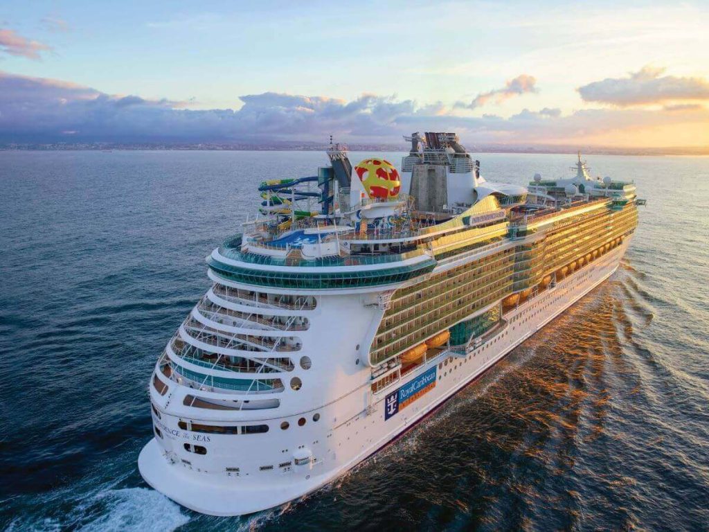 The Independence of the Seas cruise ship embarks on a voyage at sunset, its side catching the warm hues of the setting sun, with the sprawling deck activities visible and a backdrop of a distant coastline.