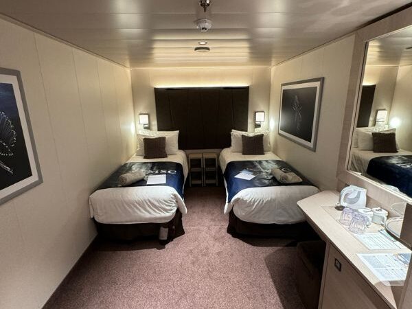 Compact and cozy inside cabin on MSC Euribia, designed with efficient use of space. The cabin features two single beds with navy blue bedding, accented with decorative pillows, flanked by nightstands and framed nautical artwork. The soft pink carpet and mirrored walls enhance the sense of space, creating a comfortable and inviting atmosphere for passengers.