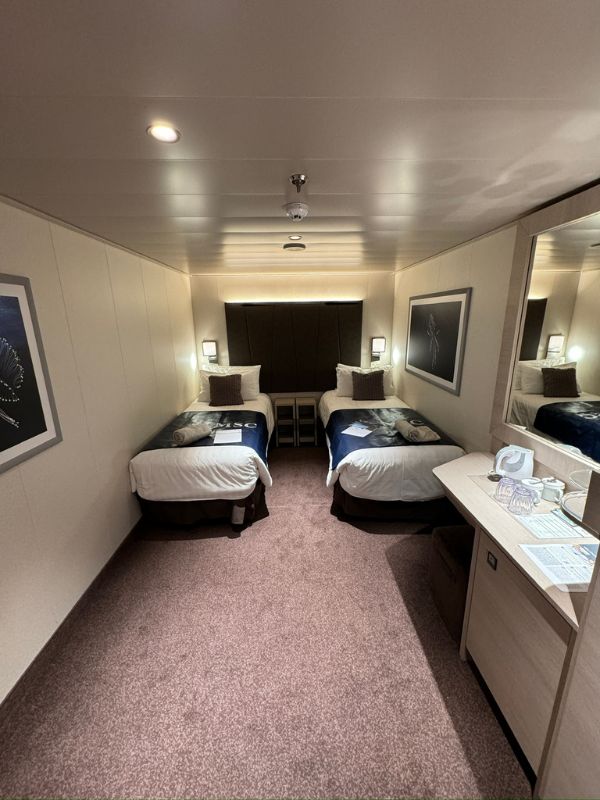 Compact and cozy inside cabin on MSC Euribia, designed with efficient use of space. The cabin features two single beds with navy blue bedding, accented with decorative pillows, flanked by nightstands and framed nautical artwork. The soft pink carpet and mirrored walls enhance the sense of space, creating a comfortable and inviting atmosphere for passengers.