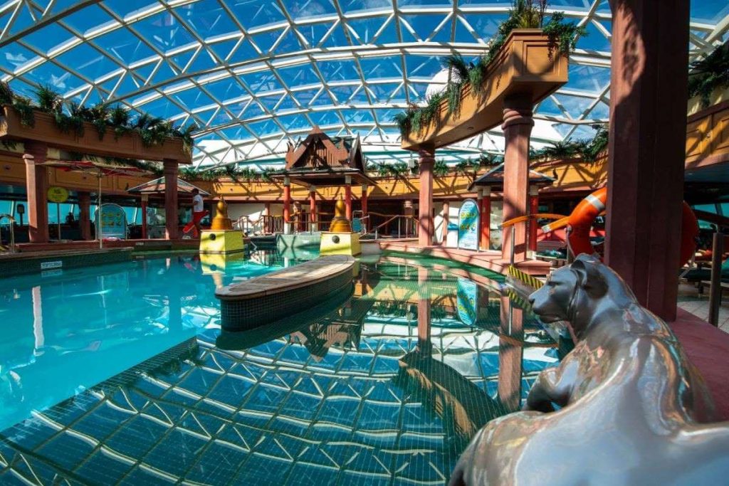 The serene indoor pool area of Royal Caribbean's Jewel of the Seas, featuring a tranquil pool, waterslide, and lounging areas under a geometric glass dome, creating a bright and airy tropical oasis.