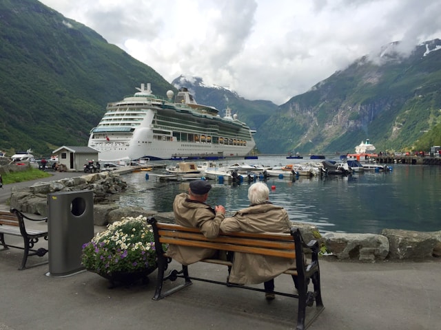 Older couple relaxing on a bench watching a Royal Caribbean cruise ship docked at port in a scenic fjord with towering mountains
