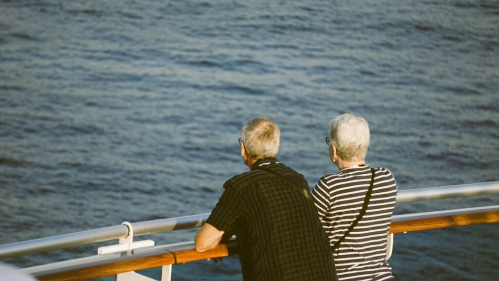 Two older adults leaning on a cruise ship railing and looking out over the ocean.