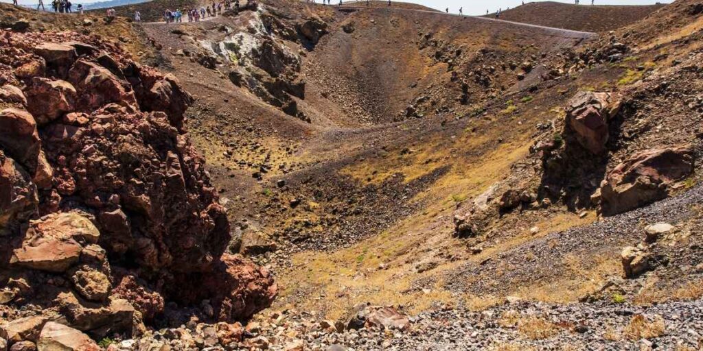 A rugged volcanic crater on Santorini's Nea Kameni island, with reddish-brown lava rocks and scattered dry grass covering the slopes. Tourists walk along the crater rim path, highlighting the area's popularity as a geological attraction within the active volcanic zone.
