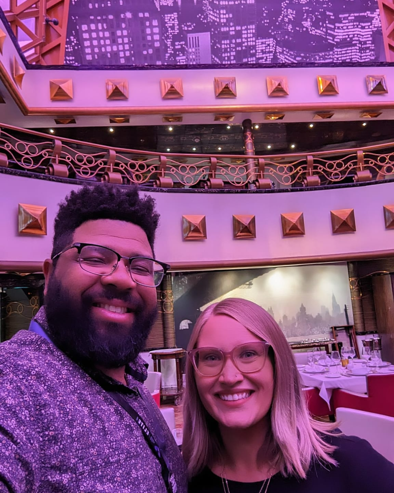 A couple smiles for a selfie inside an elegant, art deco–inspired dining room with dramatic lighting, gold geometric wall accents, and a cityscape mural in the background. Tables are set with white tablecloths and glassware, suggesting a formal dining experience, likely aboard a cruise ship.