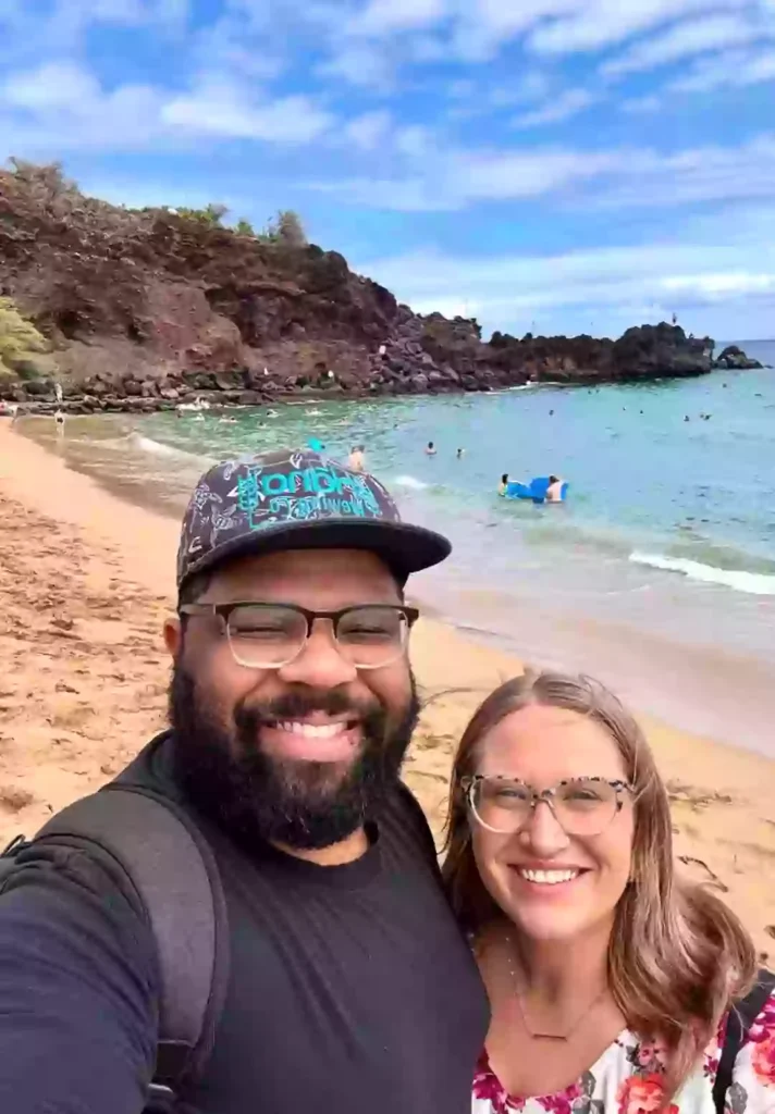A smiling couple poses for a selfie on a sandy beach with clear blue water and rocky cliffs in the background. People are swimming and relaxing in the ocean, enjoying the sunny day under a partly cloudy sky.