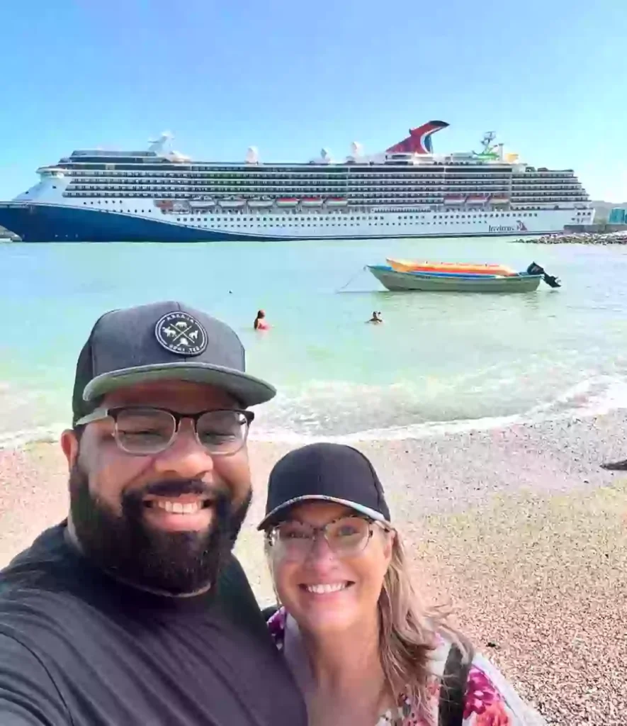 A happy couple smiles for a beachside selfie with a massive Carnival cruise ship anchored in the background. The calm turquoise water features swimmers and a small, colorful motorboat floating nearby under a clear blue sky.