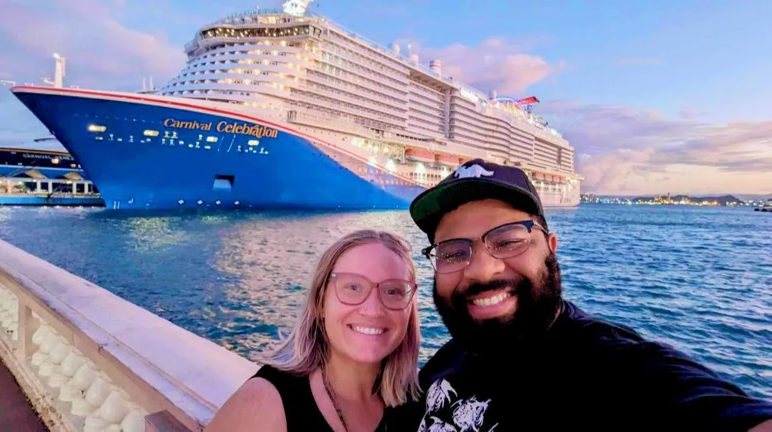 A smiling couple takes a selfie on a sunny beach with a massive Carnival cruise ship anchored in the turquoise water behind them. A small motorboat floats nearby while a few people enjoy swimming in the sea.