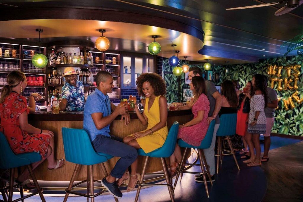A couple engaging in conversation over drinks at the vibrant Bamboo Room bar on a Royal Caribbean cruise ship, surrounded by other patrons enjoying the lively tropical-themed ambiance.