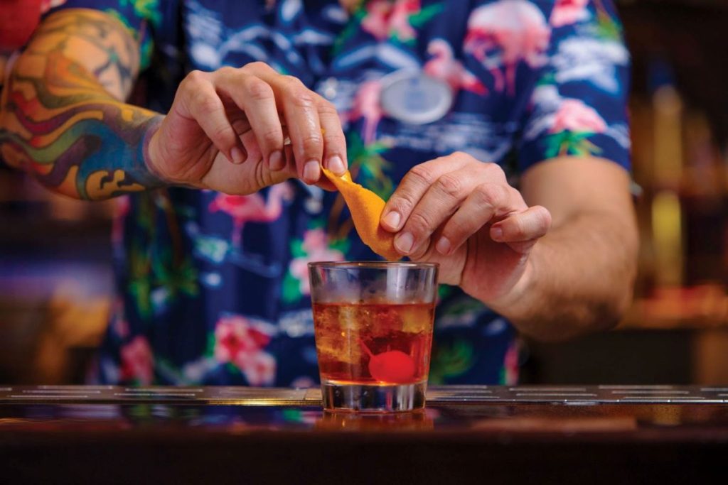Bartender with colorful tattoos preparing a cocktail at Royal Caribbean's Bamboo Room, twisting orange peel over a classic drink garnished with a cherry.