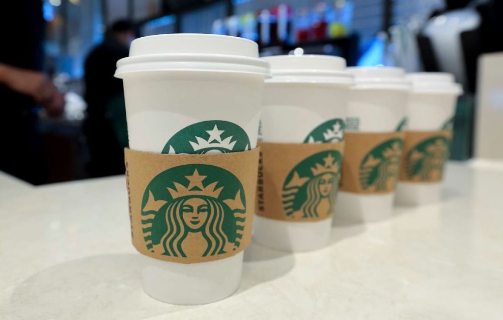 A lineup of Starbucks coffee cups with the iconic mermaid logo, arranged on a counter with a barista in the background, showcasing the partnership with Royal Caribbean.