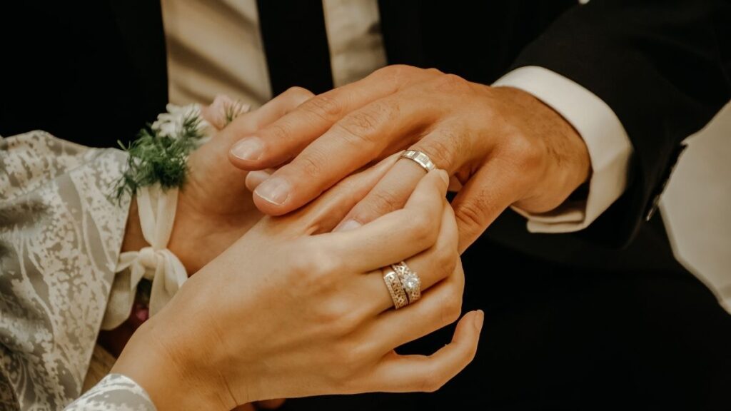 This image depicts a close-up of two hands holding each other, both adorned with wedding rings. The hands belong to a couple dressed formally, with a white floral detail visible on one hand, symbolizing a romantic or wedding moment.