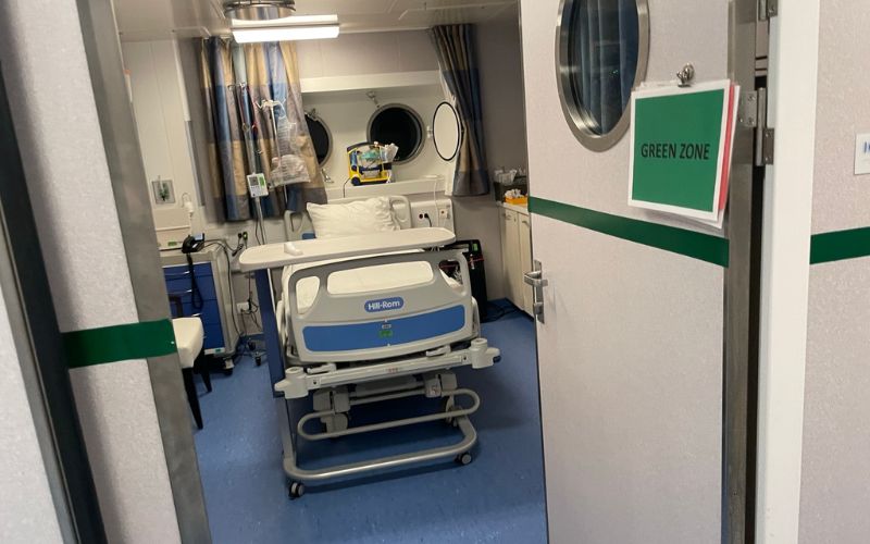 A hospital bed sits inside a clean, well-equipped cruise ship medical center room labeled “Green Zone,” with medical equipment, a porthole window, and privacy curtains visible. The space highlights onboard healthcare services available to passengers on a cruise ship.