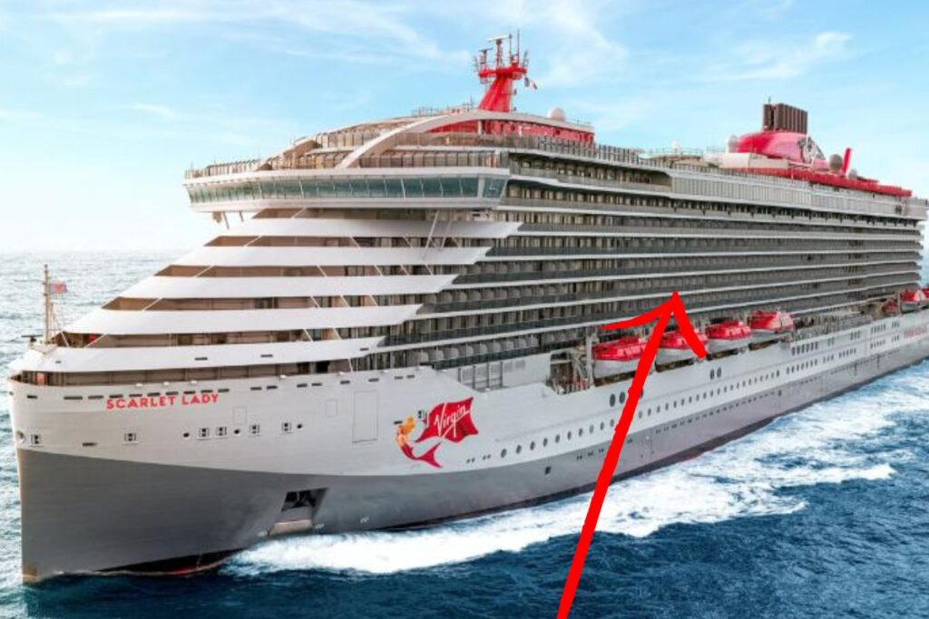 This image shows the Scarlet Lady, a Virgin Voyages cruise ship, sailing in the ocean with a focus on the middle deck. A red arrow has been added to the image, pointing directly to the midsection of the ship, highlighting the specific deck. The ship features modern, sleek architecture with several rows of balconies visible along its side, as well as the distinctive Virgin Voyages branding, including the name "Scarlet Lady" and a mermaid logo. The scene is set against a clear sky and calm seas, showcasing the ship in full motion.