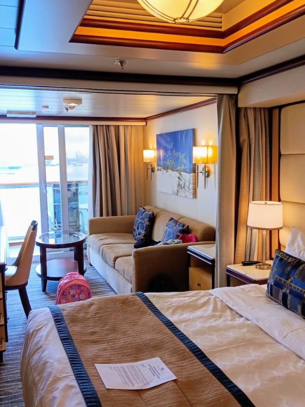 The image shows a cozy mini-suite on the Regal Princess cruise ship. The room features a large bed with neatly arranged linens, a sitting area with a sofa, and a small coffee table next to the balcony, which allows for natural light and views of the ocean. The warm, ambient lighting and stylish decor create a relaxing and inviting atmosphere, perfect for unwinding after a day at sea.