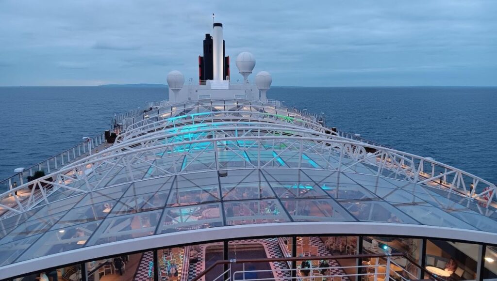 The image shows the movable roof on Cunard's Queen Anne cruise ship, providing a clear view of the ship's upper decks and the open sea. The glass-covered structure features sleek, modern design elements with sections illuminated by soft blue lighting, offering a glimpse into the dining and lounge areas below. The ship is sailing on calm waters, with the horizon stretching into the distance, while the central smokestack and radars rise above the deck. The scene highlights the ship's luxurious and innovative design, perfect for an upscale cruising experience.