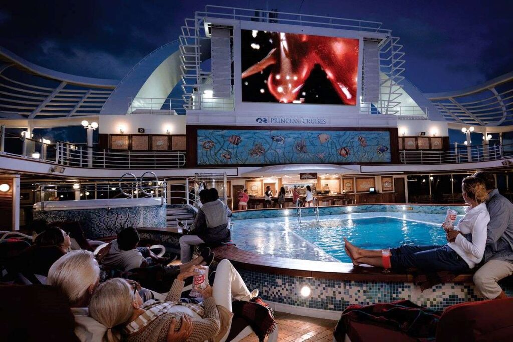 This image captures the "Movies Under the Stars" experience on a Princess cruise ship, with guests relaxing by the poolside while watching a film on a massive outdoor screen. The cozy setting features loungers and blankets for added comfort, with a beautifully lit pool and surrounding deck creating a magical evening ambiance. The illuminated screen against the night sky offers a unique and unforgettable onboard entertainment experience.
