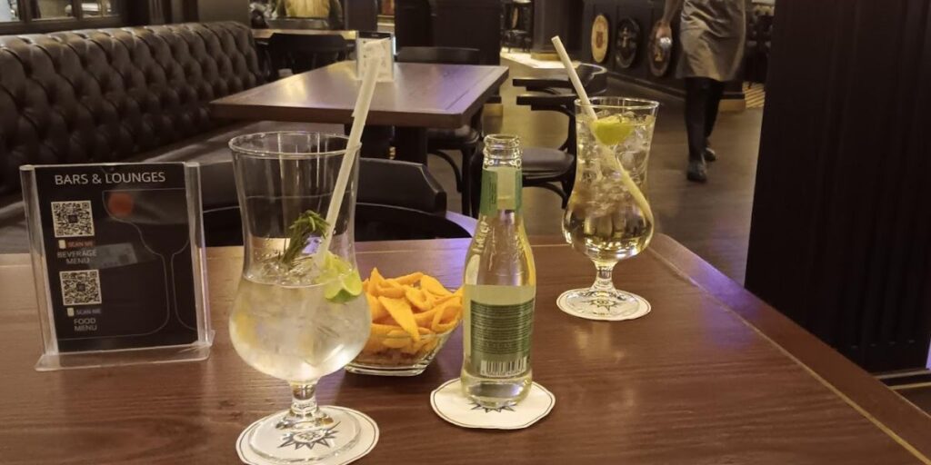 A close-up view of two refreshing cocktails on a bar table, accompanied by a bowl of snacks and a 'Bars & Lounges' menu stand, with the dark-toned interior of a lounge in the background.