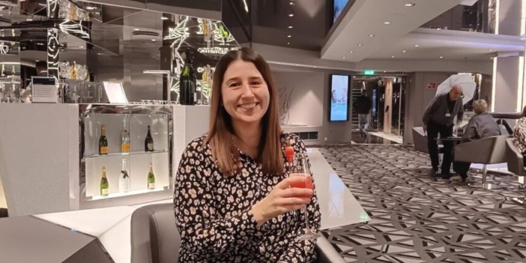 A smiling woman sitting at a bar in a modern lounge, holding a cocktail, with elegant interior decor and other patrons in the background.