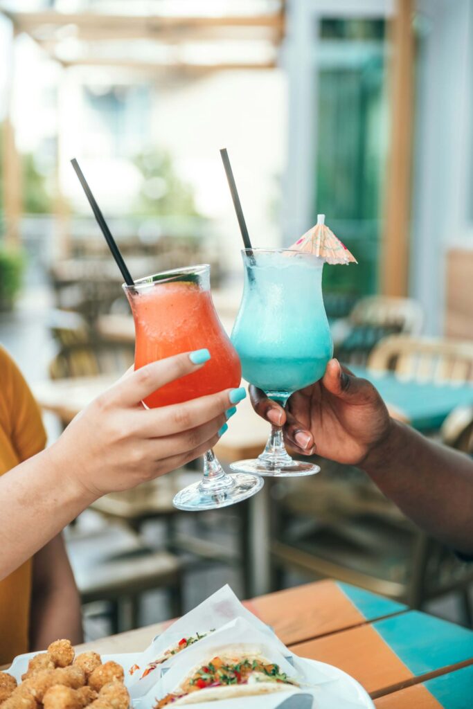 Two people clink colorful frozen cocktails—one red, one bright blue with a tiny paper umbrella—at an outdoor patio table.