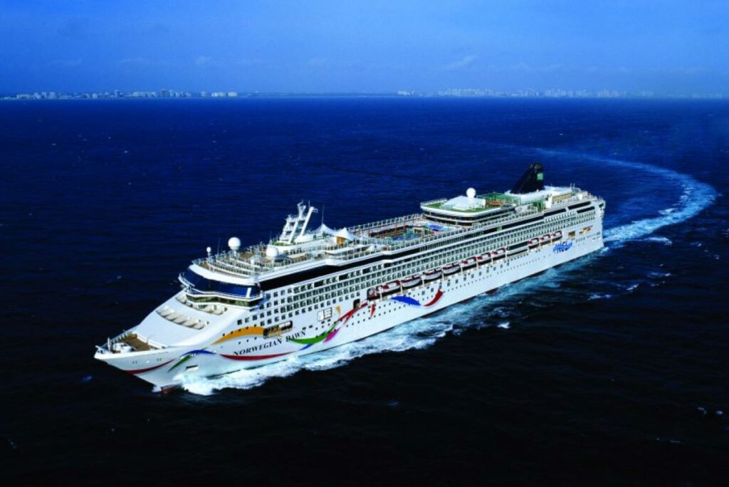 This image shows the Norwegian Dawn cruise ship cruising through deep blue waters with a distant shoreline faintly visible on the horizon. The ship’s hull is decorated with colorful wave and star-themed artwork, adding a dynamic and cheerful touch to its sleek design. The open sea and expansive sky create a serene and adventurous atmosphere.