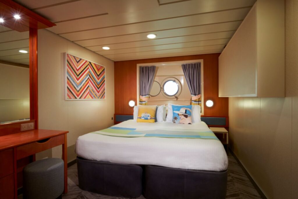 The Ocean View Porthole cabin on the Norwegian Dawn features a comfortable bed with vibrant bedding, a round porthole window offering natural light, and modern decor. The room includes a small desk and stool, colorful wall art, and ample lighting, creating a cheerful and inviting space for guests.
