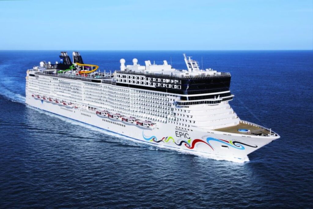 This image shows the Norwegian Epic cruise ship cruising through calm blue ocean waters under a clear, sunny sky. The ship features vibrant, wave-like artwork on its hull and boasts colorful water slides on its top deck, showcasing its family-friendly and adventurous amenities. Its large and modern design emphasizes a luxurious and dynamic cruising experience.