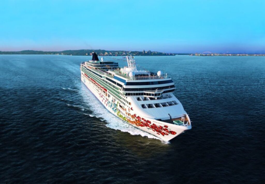 This image features the Norwegian Gem cruise ship cutting through calm ocean waters under a clear blue sky. The ship’s hull is decorated with colorful gem-themed artwork, enhancing its sleek and festive appearance. A distant shoreline is visible, adding depth to the scene and highlighting the ship’s journey toward or away from port.