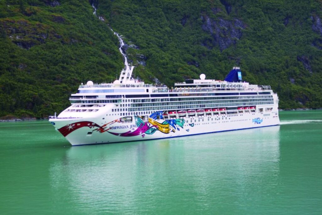 This image features the Norwegian Jewel cruise ship gliding through calm, emerald-green waters, surrounded by lush green mountains and a cascading waterfall in the background. The ship's colorful hull art adds a striking contrast to the serene natural landscape, emphasizing its grandeur and modern design. The scene showcases a picturesque and tranquil cruising experience.