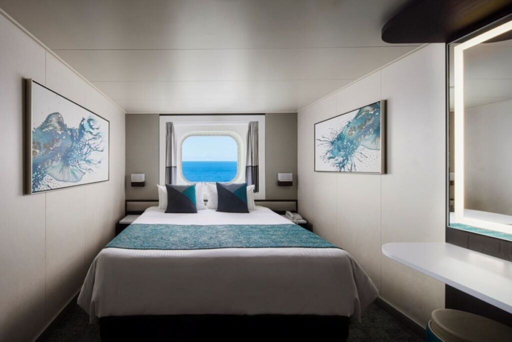 A Norwegian Jewel Family Oceanview cabin with a picture window, featuring a comfortable double bed adorned with teal and grey bedding, flanked by modern artwork and wall sconces. The window offers a beautiful view of the ocean, and a sleek vanity area is visible to the side.
