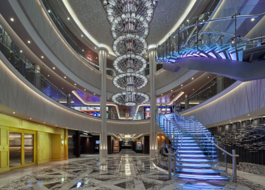 The grand atrium of the Norwegian Joy, known as Ocean Place, features a stunning, multi-tiered chandelier that illuminates the area with its intricate design. The space is complemented by a modern, illuminated staircase, sleek marble flooring, and elegant balconies that overlook the central hub, creating a luxurious and inviting atmosphere for guests.
