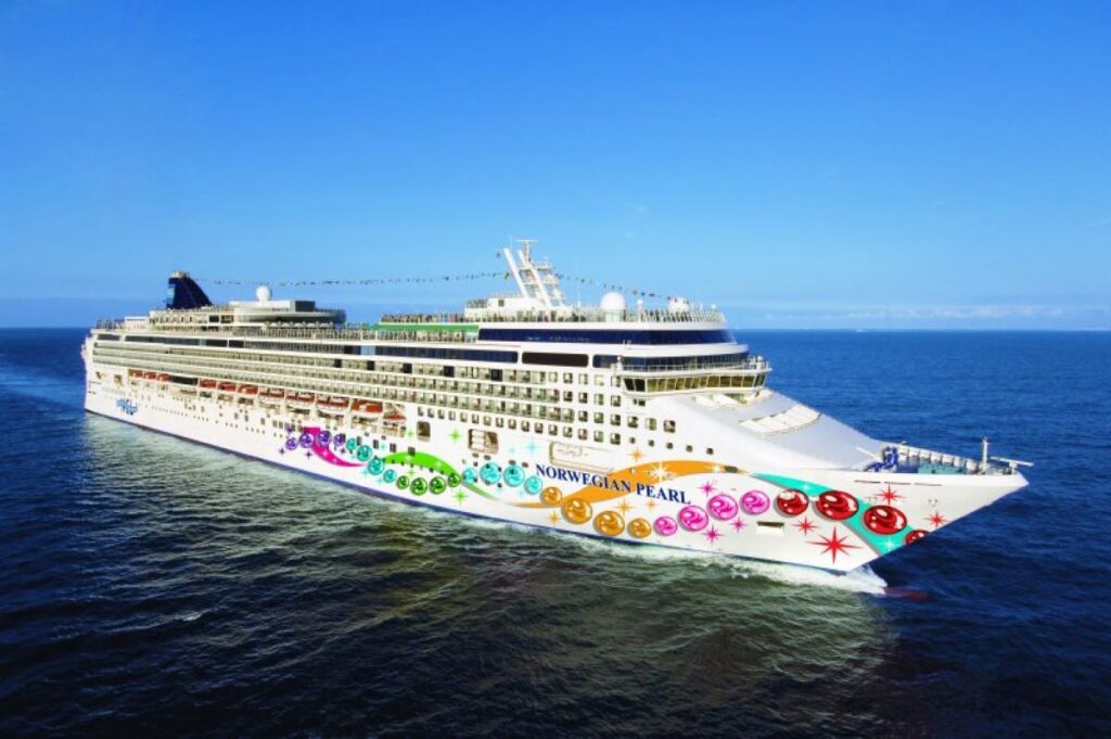 This image depicts the Norwegian Pearl cruise ship cruising through calm blue ocean waters under a clear, sunny sky. The ship is adorned with colorful pearl-themed hull art, giving it a vibrant and festive appearance. Its sleek design and expansive decks reflect the luxury and grandeur of this maritime vessel.