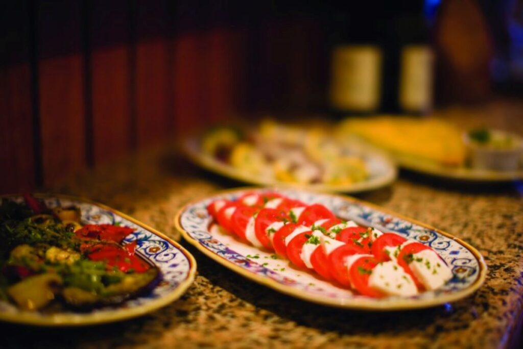 This image showcases dishes from La Cucina, a dining venue on an NCL ship. The focus is on a plate of fresh Caprese salad, featuring alternating slices of ripe tomatoes and mozzarella, garnished with fresh herbs. The setting is warm and inviting, with intricate ceramic plate designs and additional Italian-inspired dishes blurred in the background, enhancing the ambiance of fine dining at sea.