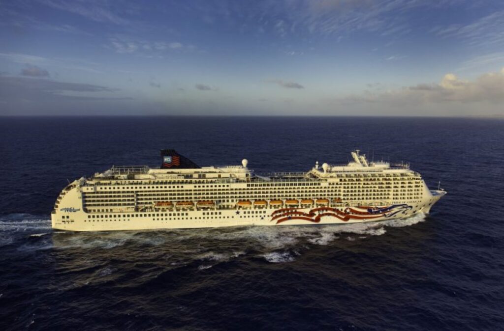 This image features the NCL Pride of America cruise ship navigating through the vast ocean under a partly cloudy sky during what appears to be golden hour. The ship's hull displays a patriotic design with red, white, and blue waves and a U.S. flag emblem, reflecting its American theme. The serene open-water setting emphasizes the ship's grandeur and its connection to leisurely exploration.