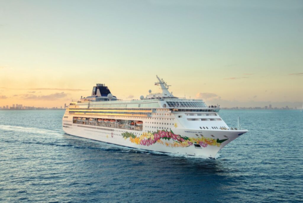 This image features the Norwegian Sky cruise ship cruising through calm waters at sunset, with a distant city skyline visible on the horizon. The ship’s hull is decorated with vibrant floral artwork, adding a touch of elegance and whimsy to its sleek design. The golden hues of the sky and water create a serene and picturesque atmosphere.