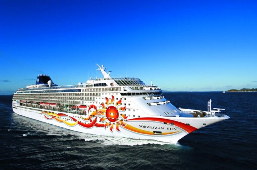 This image showcases the Norwegian Sun cruise ship sailing through deep blue waters under a clear, vibrant sky. The ship’s hull features bright sun-themed artwork, reflecting its warm and inviting design. The scene exudes a sense of adventure and relaxation, with a distant shoreline visible in the background.