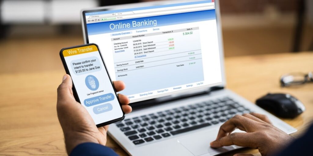 Person using online banking with a laptop displaying account balances and transactions, while holding a smartphone showing a wire transfer screen to approve a $120 transfer to Jane Doe using fingerprint authentication.