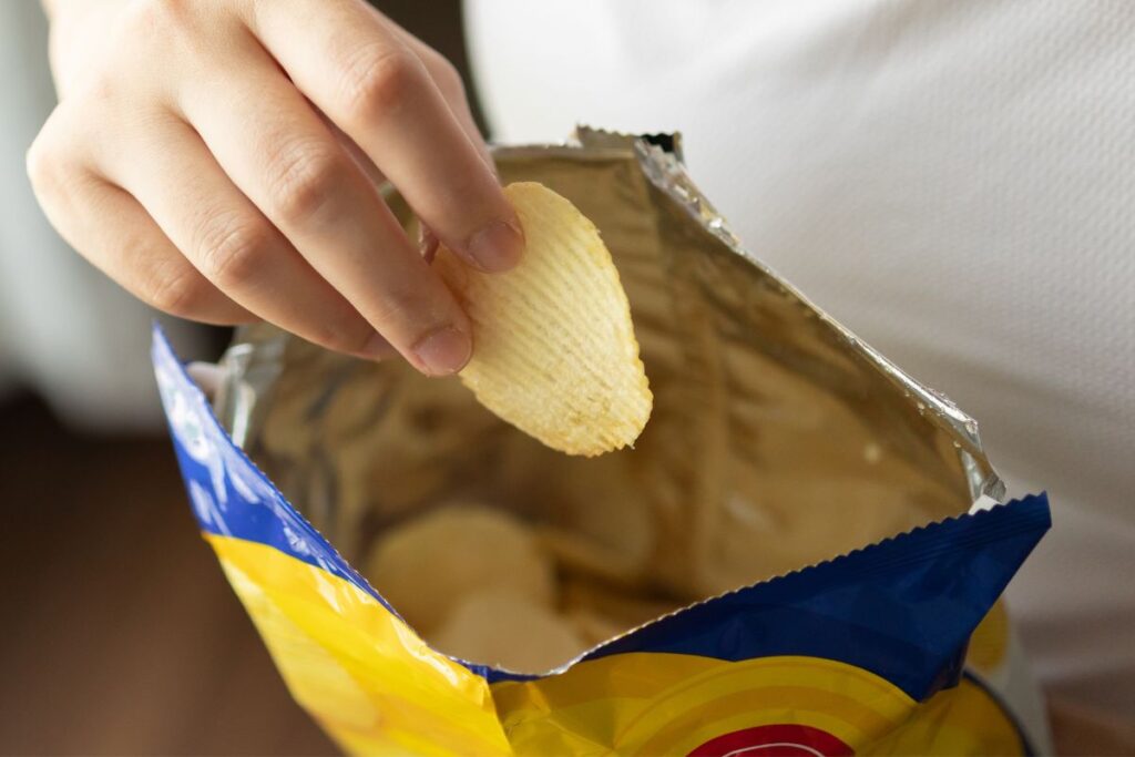 The image depicts a hand reaching into an open packet of crisps, holding a single ridged chip. The crisps are in a foil-lined bag with vibrant blue and yellow packaging, suggesting a casual snack moment.