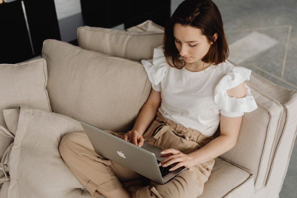 Woman in a white ruffled blouse and beige pants focused on cancelling her Carnival cruise on her MacBook while comfortably seated on a beige couch.