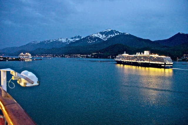 Cruise ship sailing past snow-capped mountains at dusk in Alaska, viewed from a ship’s deck over calm water.