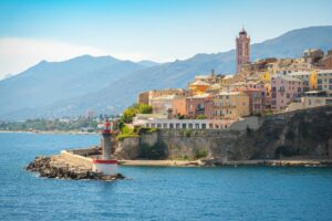 Colorful hillside buildings in Bastia, Corsica overlooking the sea, with a lighthouse on the breakwater and mountains in the background.