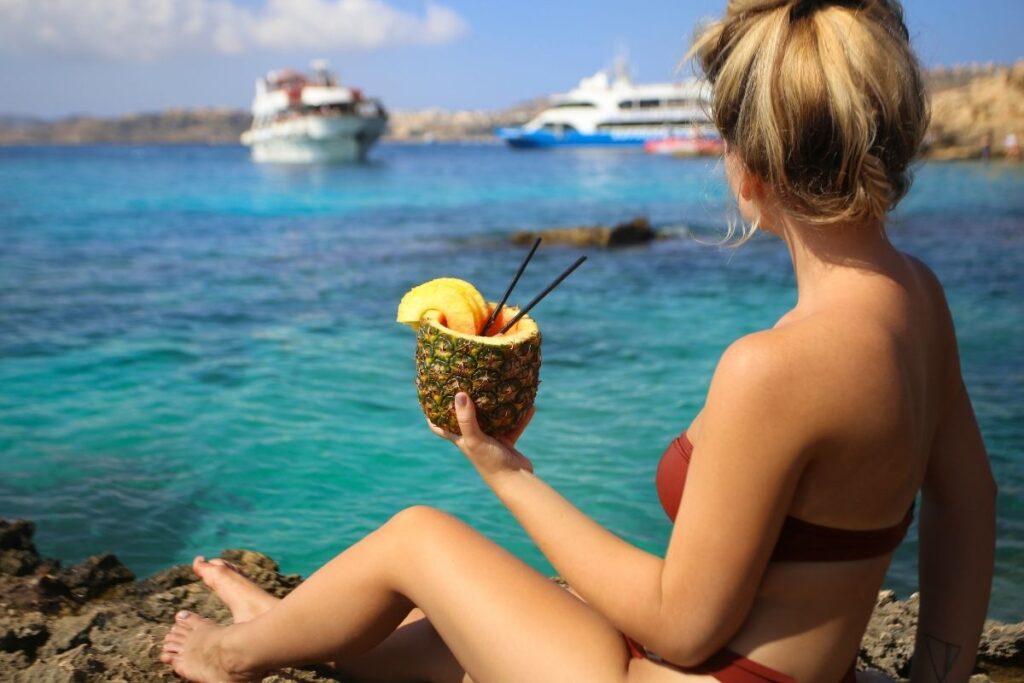 This image shows a woman in a bikini sitting on rocky terrain near crystal-clear turquoise waters, holding a tropical drink served in a pineapple. In the background, cruise ships are anchored, adding a touch of luxury and adventure to the serene coastal scene. The image captures the essence of relaxation and indulgence during a tropical getaway.