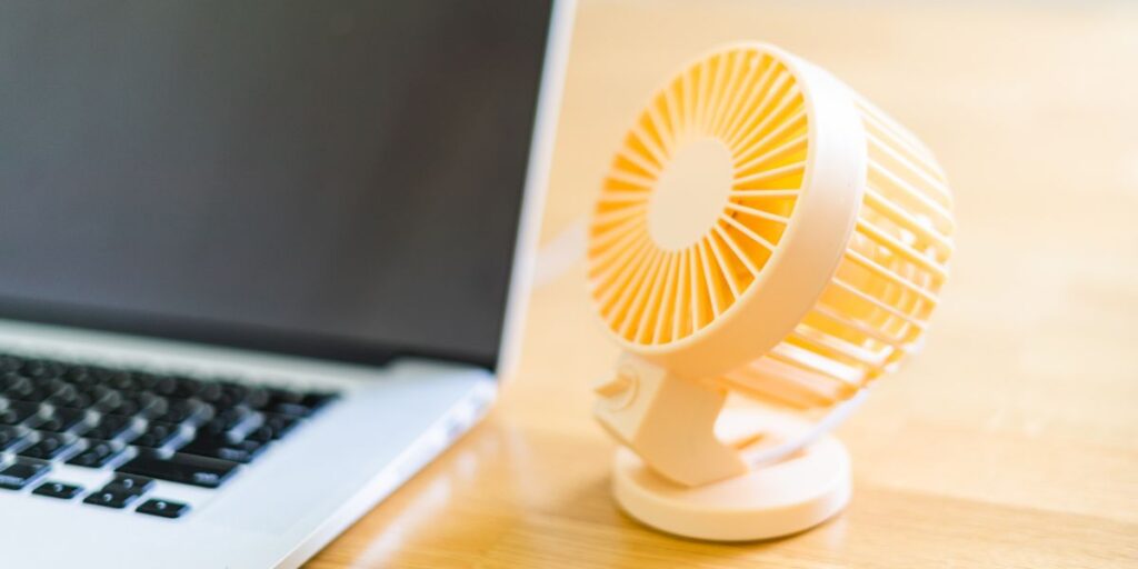A small, yellow portable fan positioned next to an open laptop on a wooden surface, suggesting a comfortable work environment.